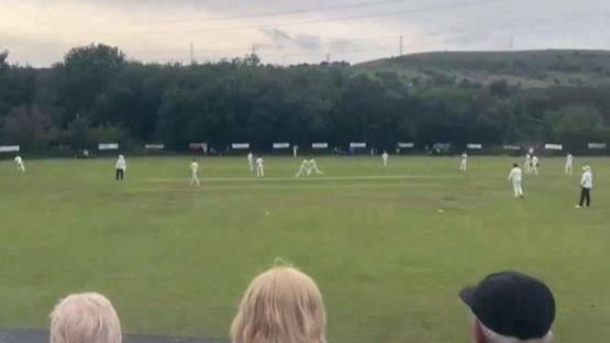 Wide view of teams in white cricket kit playing on a green pitch surrounded by trees and hills. Three seated specatators can be seen at the bottom of the image watching the game.