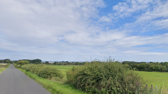 A greenfield site with a rural road, bushes and grass to one site