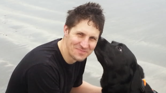 A man is on a beach with a black dog licking the side of his face. The man is wearing a black T shirt.