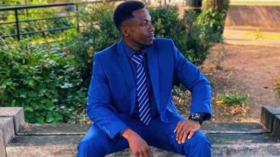 Allen Tejan sitting on a wooden bench outside. He is smartly dressed in a bright blue suit and striped blue tie.