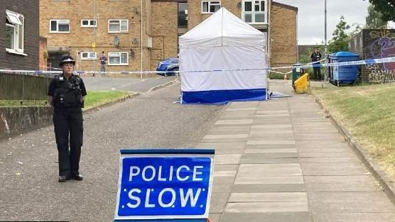 Police officer stands close to the cordon and a Police Slow sign, with a white forensic tent in the background