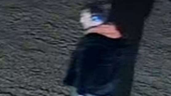 A blurred CCTV still image of a man with light hair. He is wearing blue headphones, a black jacket and an orange hoodie.