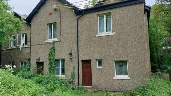 Holly Lodge before the fire. The building is two storeys high and looks more like a row of cottages, with two wooden front doors. The nearest part of the house is clad in small stones while to the left the building has larger stonework. There are a series of pitched roofs and the garden is very overgrown.