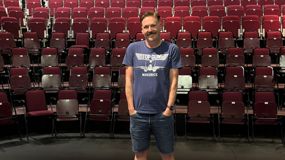 Shows a man with a moustache and a blue and white Top Gun - Maverick t-shirt with his hands in the pockets of denim shorts standing in an empty theatre of red seats
