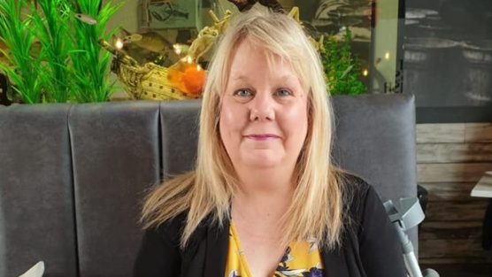A woman, in her 50s, with long blonde hair. She is wearing a floral yellow dress under a black cardigan and is smiling