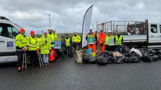A dozen people in yellow and orange hi-vis clothing stand either side of two white pick-ups with around 20 black bags of rubbish in the foreground