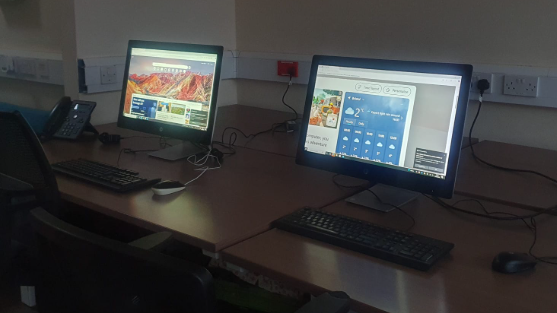 Image shows two desktop computers lit up in a dark room. The desktops are on two wooden tables with desk chairs. 