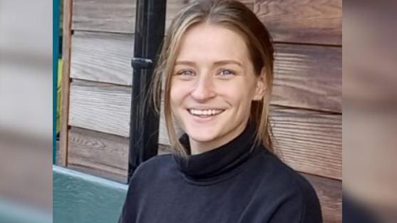 A young woman with light brown hair in a ponytail, wearing a black turtle neck and blue jeans sits on a bench. She is smiling at the camera.