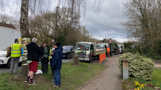 A group of people stand near a tree with tape wrapped around its trunk. They stand on a strip of grass on a residential road. Several vans are parked down the road.