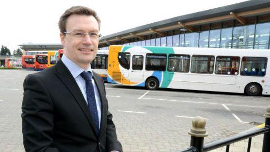 Stagecoach East Midlands Managing Director Matt Cranwell standing to the left of the photo. He is middle-aged wearing a black blazer, light blue shirt and navy tie with white spots. He is smiling, has dark hair and is wearing silver thin framed glasses. He is in a coach park, with a row of yellow, white and turquoise single decker buses behind him. 