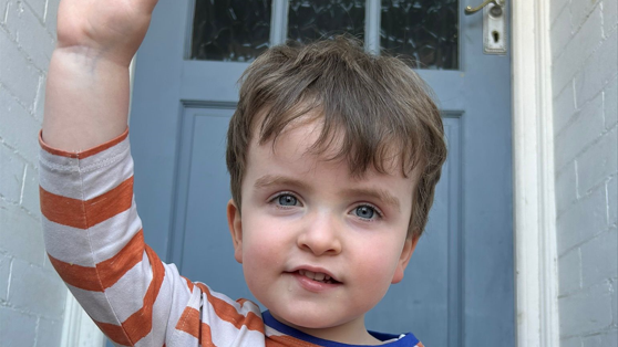 Charlie is sat down by a front door waving. He has an orange stripy top on. He has short brown hair.