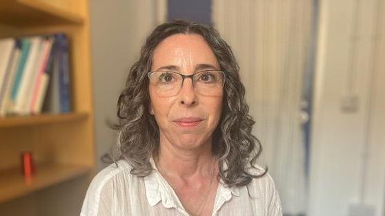 Prof Abigail Fraser, wearing white top and glasses, while standing in an office room with a book shelf in the background.