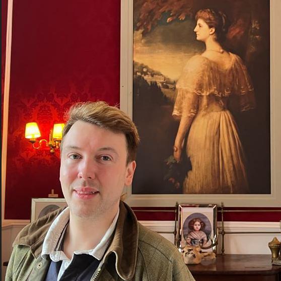 Mark Baker, in a green stands under a painting of a woman on a red wall smiling