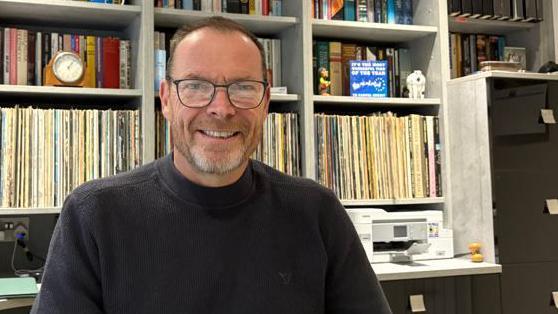 Image of Phil who is sat in front of a bookcase full of books. He is wearing a navy jumper and is wearing glasses.