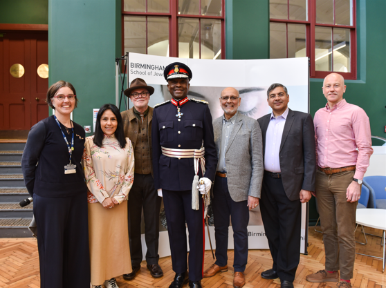 A group of men and women smile at the camera, while standing in front of a white board saying Birmingham School of Jewellery. There are two women and five men in the group. The man in the middle wears a blue military-looking uniform and black cap.