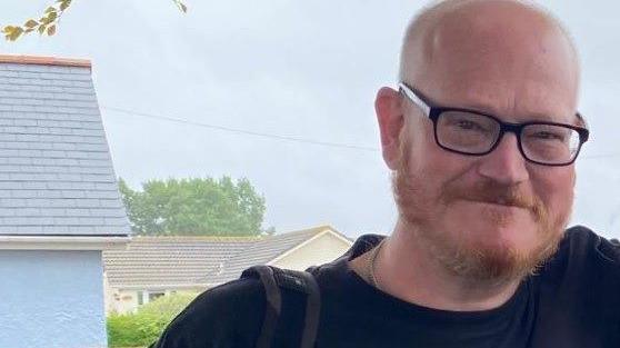 Ian Airlie is seen with glasses and a beard is smiling outdoors, with houses and trees visible in the background on a cloudy day.