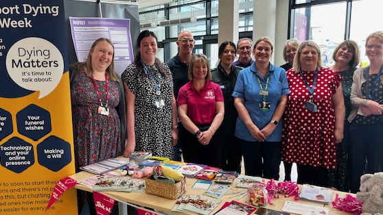Nine women and two men, many wearing name badges, stand behind a table covered in leaflets and pens. A pull-up stand reads "Dying Matters, it's time to talk about it".
