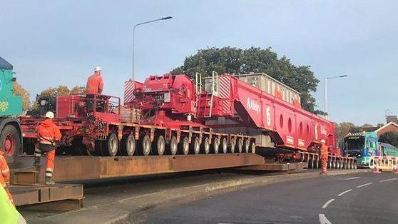 A general view of an abnormal load on a road. Highways workers in orange hi-vis  coats and trousers stand near or on the abnormal load. 