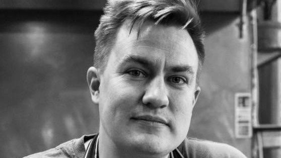 A close-up black-and-white image of Adam Armstrong, in a kitchen. He has short hair and is smiling.