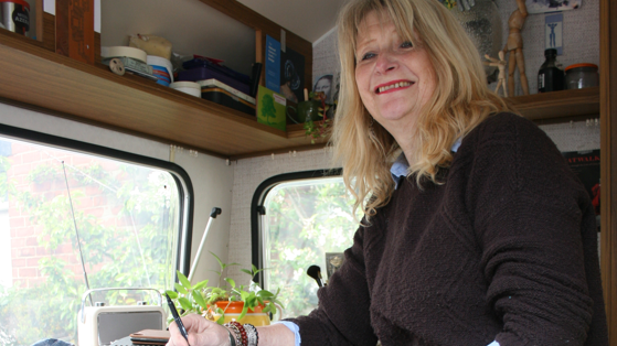 Angela Sandwith is smiling as she writes in her studio. Her blonde hair is just over her shoulders and she has blue eyes. She is wearing a brown jumper over a blue shirt and has red lipstick on. Her studio is small and she is surrounded by books and plants.