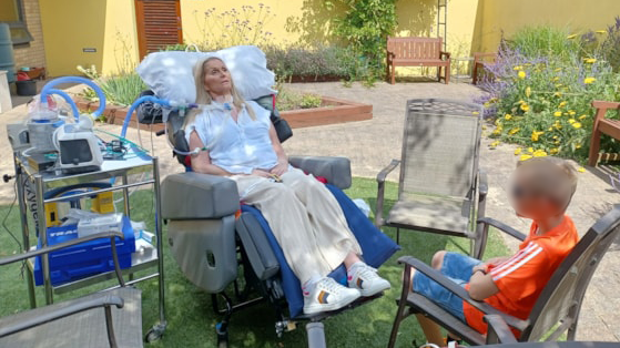 Catherine lying in a medical bed in a garden. She is connected to medical equipment. A young boy is sat by the bed.