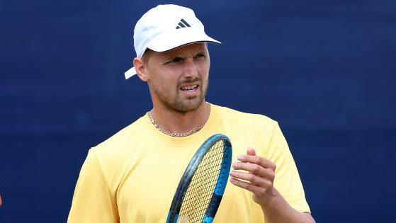 Lloyd Glasspool holding his racquet wearing a yellow t-shirt and a white Adidas cap