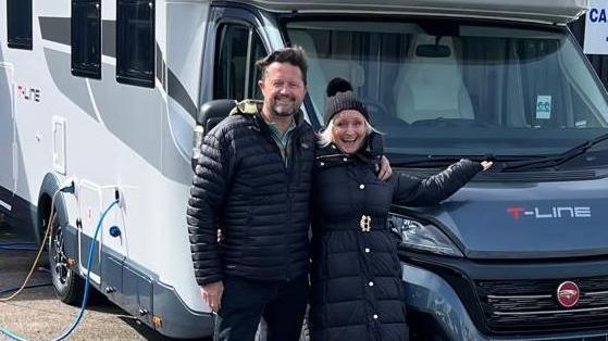 A man and woman stood outside in front of their motorhome. The man has black hair, swept to one side. He is in a puffer jacket and has camel coloured trainers on. He has his left arm around the woman's shoulder. She is wearing a black bobble hat and smiling. She has blonde hair and is wearing a long black puffer coat. 