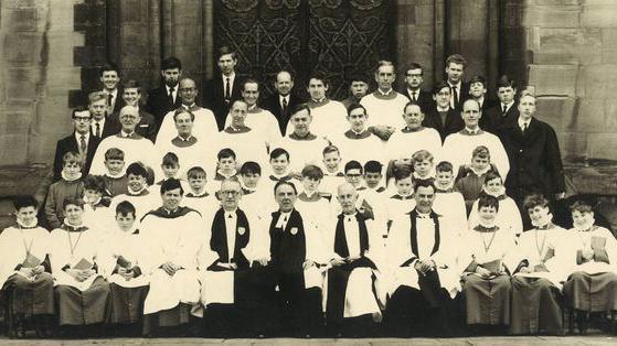 A black and white photo of a group mixed aged males dressed in white gowns and standing in rows.