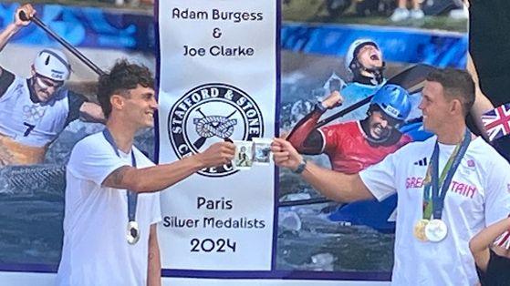 Two men wearing Olympic medals around their necks are clinking mugs together in "cheers". There is a banner behind them, with photos of them in their canoes. The banner reads "Adam Burgess and Joe Clarke. Paris silver medallists 2024." 