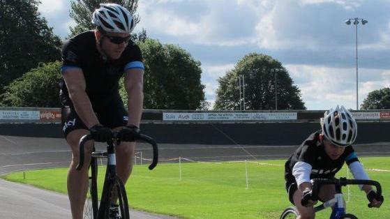 Archie Atkinson is cycling next to Sir Chris Hoy on an outside cycling track. They are both wearing helmets and cycling gear.