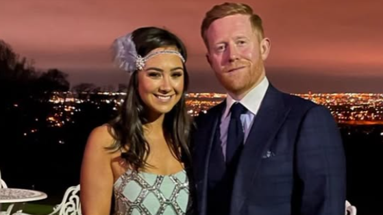 A man and a woman dressed smartly smiling at the camera. The woman has a feather bow in her hair and the ginger man is wearing a suit and tie