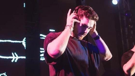 A man in a dark T-shirt holding headphones up to his head. The background is black with white lines behind him.