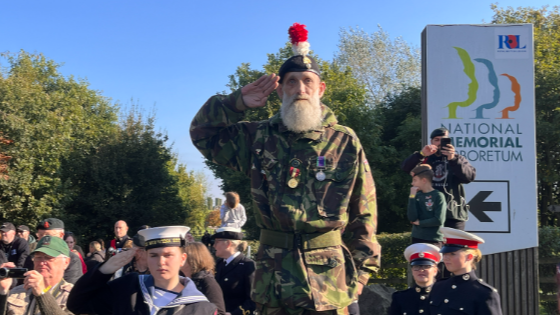 A man in camouflage, wearing a beret with a red and white plume, salutes, with others in military uniform stood around him 