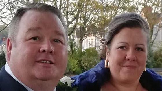 Christopher Mills a Michelle Mills ar ddiwrnod eu priodas. Christopher Mills mewn siwt ddu a Michelle Mills yn gwisgo sgarff las a chlustlysau aur.