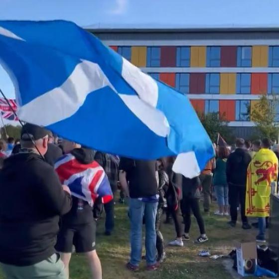 A protest outside the Hampton by Hilton hotel in Westhill, Aberdeenshire. A man carries a large saltire in the foreground. Another man is draped in a union jack, and a third has the lion rampant around his shoulders. All have their backs to the camera.