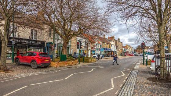 Westgate in Guisborough. Cobbles line the road, while tall trees are growing  in planters along the road. A jewellers is among a number of shops on the high street.