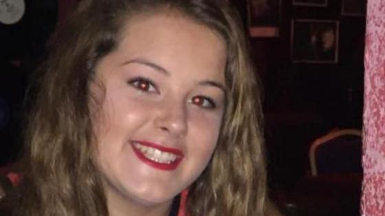 Emily smiles at the camera. She has long wavy brown hair and red lipstick on.