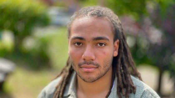 A man with dreadlocked hair looks straight at the camera. He is standing in an bright, green, outdoor setting which has been blurred.