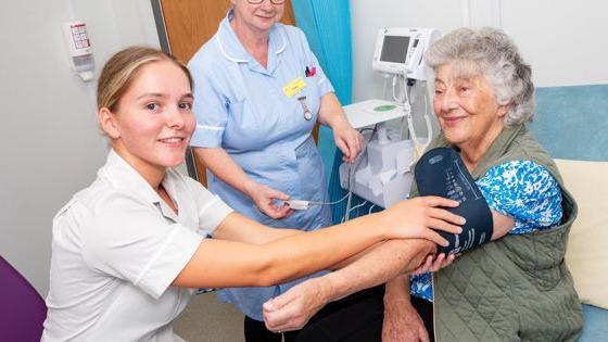 An older woman is sitting down, she has curly grey hair and her arm is outstretched. It has a blood pressure monitor on it. A younger woman sits next to her, in a white shirt. She is holding the blood pressure monitor. Another woman is behind them in nurses scrubs, standing next to a white medical machine