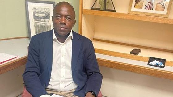 Man wears a white shirt and navy jacket sits on an armchair with a clipboard on his lap. He has framed photographs on shelving behind him.