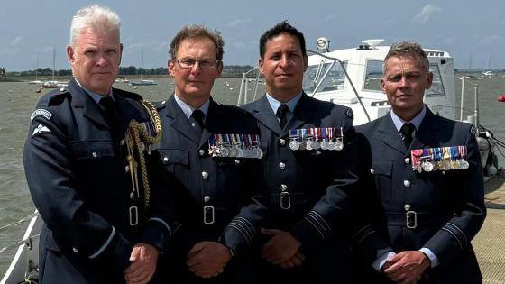 Matt Stowers, Andy Calame, Mike Dewar and Mark Jacklin are all wearing Armed Forces uniform, and have lots of medals pinned to their jackets. A white boat and the water is behind them.