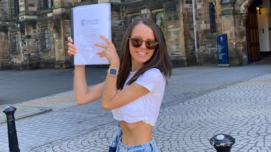 Hayley Morrison holds her dissertation up in front of the University of Glasgow main building. She wears a white cropped T-shirt, sunglasses and blue jeans. She smiles at the camera.