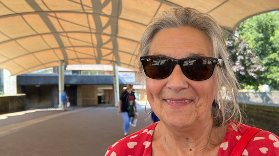 A woman with tied-back grey hair and sunglasses. She is wearing large hoop earrings and a red dress with love hearts on it and a red bag over her shoulder. Behind her is a flowering tree and people walking.