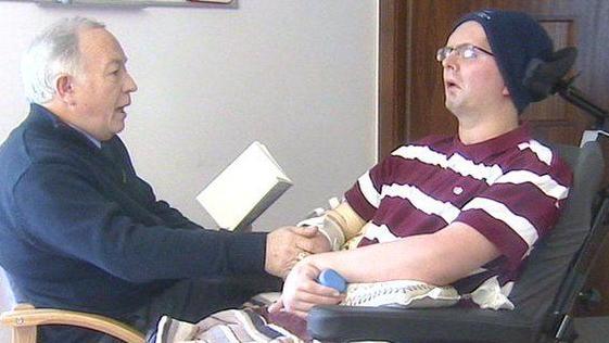 Ann elderly man is reading a book to a man who is in a wheelchair. They are holding hands. The older man is wearing a navy jumper. The younger man is wearing a stripe polo shirt, it is burgundy and white. 
