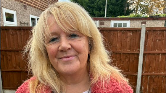 Heather Matuozzo has shoulder-length blonde hair and is wearing a pink top and is smiling at the camera. She is standing in a garden with a brown fence behind her.