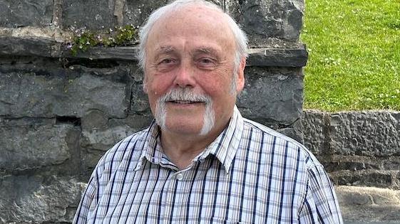 Tony Brown, a man with white hair and a long moustache. He is wearing a white shirt with dark grey lines, and is smiling as he standing in front of a stone wall that is part of Castle Rushen on the edge of Market Square.