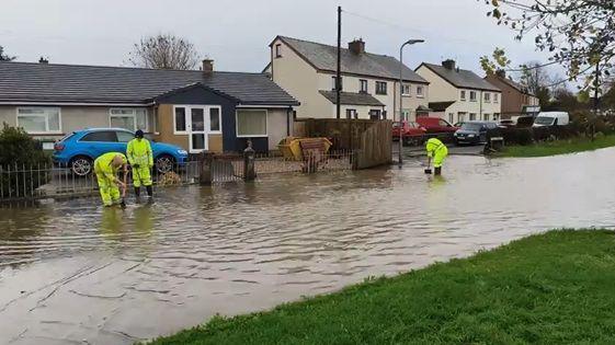 Three people in high-visibility clothing work to clear drains as a road is flooded in the village of Thursby. There is muddy water which is reaching homes.Â