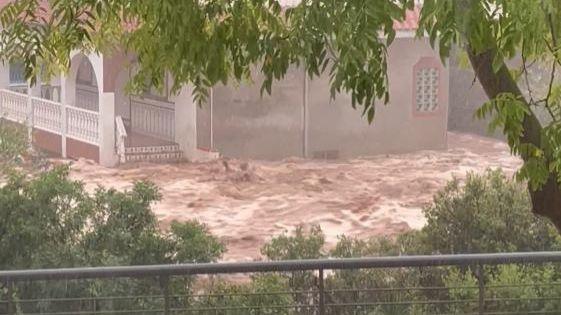 Brown floodwaters surge past a house in La Rapita, Spain, with a tree and some plants visible from an elevated position in the foreground