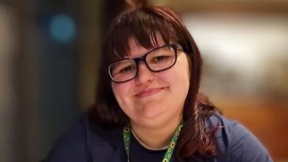 Image of Sophie. She has brown hair is wearing black-rimmed glasses. She is wearing a green lanyard around her neck.