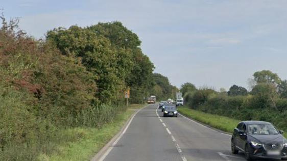 Cars travelling down an A-road in the countryside on a sunny day. The road is lined by trees to the left side and a hedge to the right side.